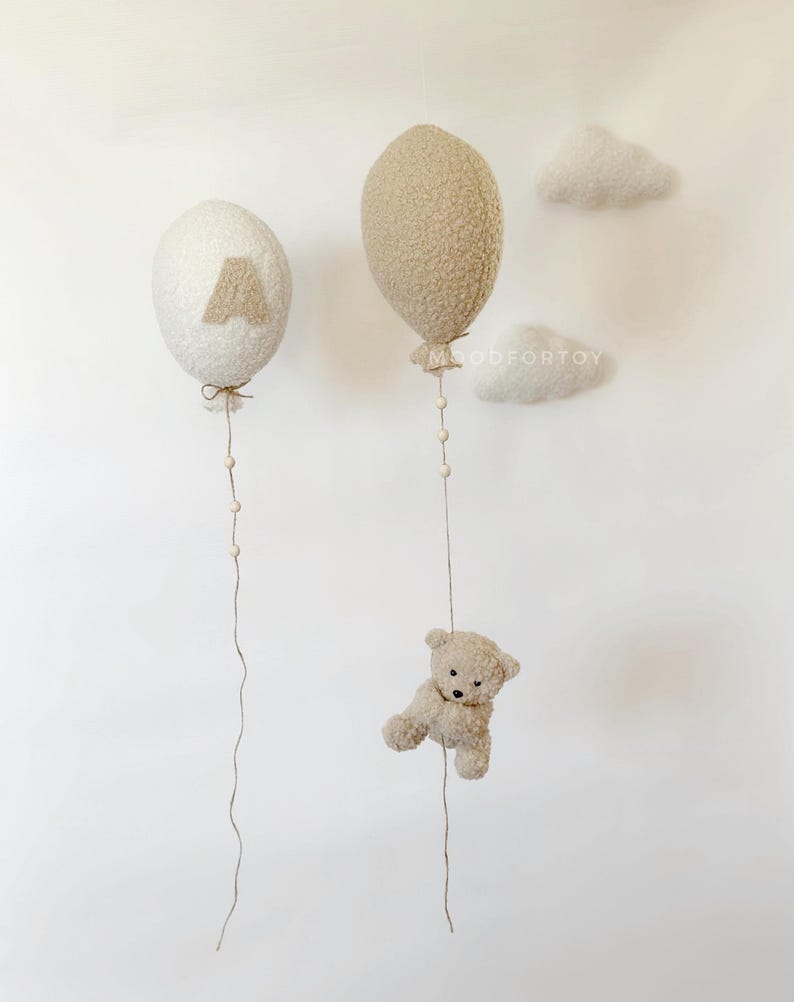 K&ouml;nnte beinhalten: Zwei flauschige, cremefarbene Ballons mit einem braunen Buchstaben 'A' auf einem Ballon und einem braunen Teddyb&auml;r, der am Boden des anderen Ballons h&auml;ngt. Die Ballons h&auml;ngen an einer wei&szlig;en Wand mit zwei wei&szlig;en, flauschigen Wolken.