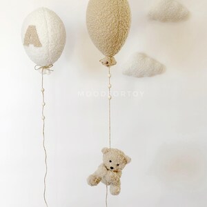K&ouml;nnte beinhalten: Zwei flauschige wei&szlig;e und beigefarbene Ballons mit Schnur und Holzperlen, die von ihnen h&auml;ngen. Ein Teddyb&auml;r h&auml;ngt an dem beigen Ballon. Die Ballons h&auml;ngen an einer wei&szlig;en Wand mit zwei wei&szlig;en, flauschigen Wolken.