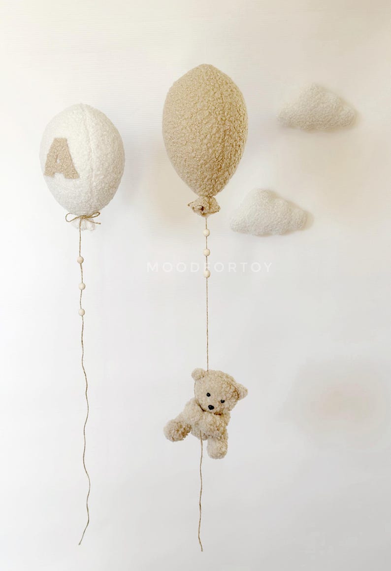 K&ouml;nnte beinhalten: Zwei flauschige wei&szlig;e und beigefarbene Ballons mit Schnur und Holzperlen, die von ihnen h&auml;ngen. Ein Teddyb&auml;r h&auml;ngt an dem beigen Ballon. Die Ballons h&auml;ngen an einer wei&szlig;en Wand mit zwei wei&szlig;en, flauschigen Wolken.