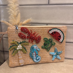 May include: A beige woven clutch bag with a zip, decorated with beaded sea-themed motifs. Includes a red lobster, a green turtle, a blue seahorse, a starfish, a palm tree, and a watermelon slice.