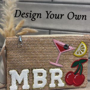 May include: A tan straw clutch bag with a zipper closure. The bag has white felt letters that spell "MBR" and is decorated with colourful embroidered patches of a cocktail glass, a lemon slice, and cherries.