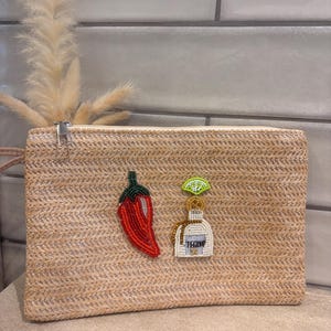 May include: A rectangular straw clutch bag featuring a beaded red chilli pepper and a beaded white tequila bottle with a lime wedge on top. The bag has a zip closure at the top and a woven texture throughout the exterior surface.