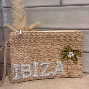 May include: A rectangular straw clutch bag with a wrist strap, featuring the word IBIZA in white pearl-embellished letters and a beaded palm tree design with green leaves and a tan trunk, secured with a silver-toned metal zip closure.