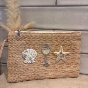 May include: A rectangular straw clutch bag featuring three beaded appliques on the front: a white seashell, a wine glass with silver and cream beads, and a cream-coloured starfish. The bag has a silver zipper closure and a wrist strap.