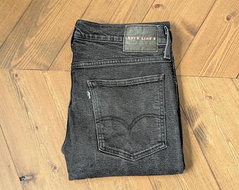 May include: Folded black denim jeans with a leather Levi's Line 8 label. The jeans are folded neatly and are set against a light brown wooden background.