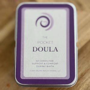 May include: A rectangular tin with a purple and white design. The text "THE POCKET DOULA" is visible, along with "50 CARDS FOR SUPPORT & COMFORT DURING BIRTH". The tin is on a wooden surface.
