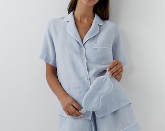 May include: A light blue pajama set consisting of a short-sleeved button-down shirt with a collar, a chest pocket, and matching shorts. Both pieces feature white piping trim. A matching light blue sleep mask with white piping is also included.