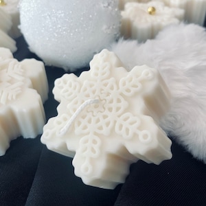 Peut inclure: Gros plan sur des bougies blanches en forme de flocon de neige avec un motif texturé. Une sphère blanche recouverte de paillettes et une matière blanche duveteuse sont également visibles. Les bougies ont une mèche et sont posées sur un fond noir.