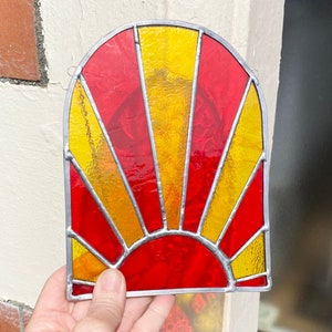 May include: A stained glass sunburst window hanging in a window frame. The sunburst is made of red and yellow glass with a silver lead frame.