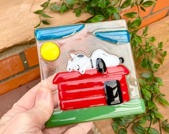 May include: A square fused glass art piece featuring a white dog with black spots sleeping on top of a bright red doghouse. The scene includes a yellow sun, two blue clouds, and a green base, all set against a clear, textured glass background.