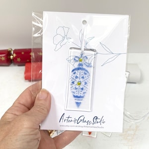 Può includere: Un ornamento di vetro blu e bianco con un motivo a fiocco di neve e accenti gialli. L'ornamento è appeso a una corda bianca ed è confezionato in un sacchetto di plastica trasparente con un cartoncino bianco che dice "Artemis Glass Studio" e un indirizzo del sito web.