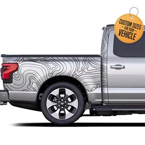 May include: A silver pickup truck with a topographic map design on the side. The map is black and white, covering the truck bed and part of the cab. A yellow tag reads "CUSTOM SIZES FOR YOUR VEHICLE".