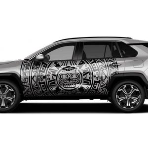 Puede incluir: SUV plateado con un diseño tribal en blanco y negro en el lateral. El diseño presenta un motivo central en forma de rostro rodeado de patrones geométricos. El vehículo tiene ruedas negras y un diseño elegante y moderno.