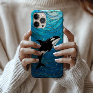 May include: A blue phone case with a black and white orca whale design. The case has a wavy pattern with gold accents. The phone case is held by a person wearing a cream-coloured sweater.