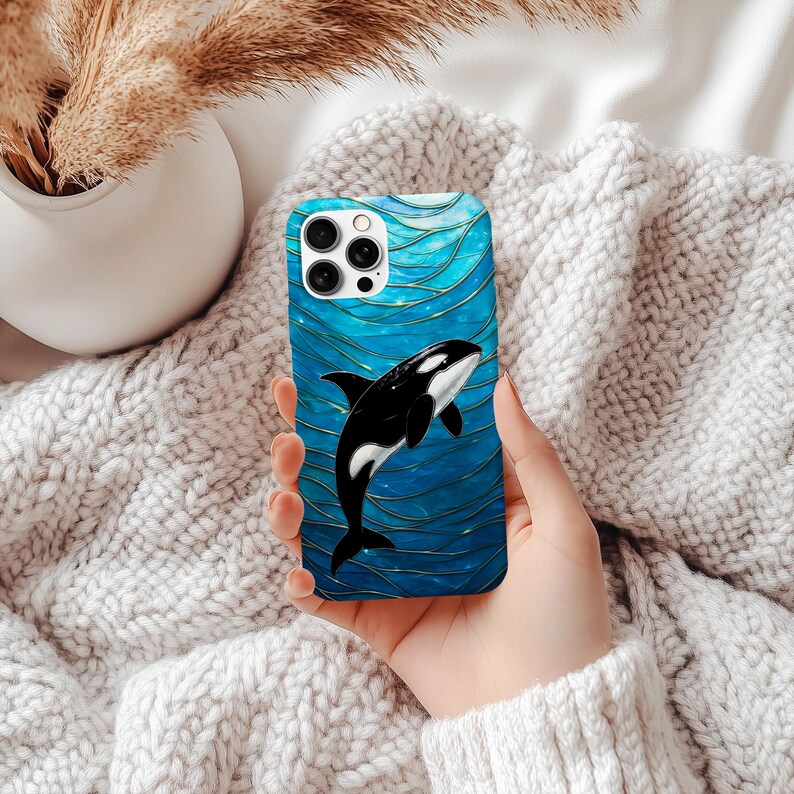 May include: A blue phone case with a black and white orca whale design. The case has a glossy finish and a wave-like pattern in shades of blue and gold. The phone case is held in a person's hand.