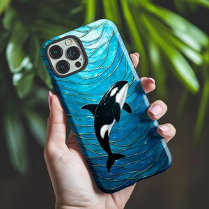 May include: A blue and gold phone case with a black and white orca whale design. The case has a glossy finish and a wave-like pattern. The phone case is held in a person's hand.