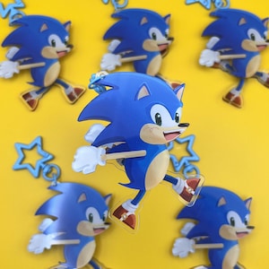 Könnte beinhalten: Eine Sammlung blauer Sonic the Hedgehog-Schlüsselanhänger mit weißen Handschuhen und roten Schuhen, vor gelbem Hintergrund. Jeder Schlüsselanhänger zeigt die Figur in einer Laufpose. Sternförmige Charms sind ebenfalls vorhanden.