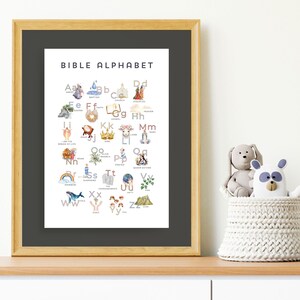 May include: A framed print titled "BIBLE ALPHABET" with watercolor illustrations for each letter. The artwork features religious imagery, including angels, a church, and a rainbow. The frame is light brown, and the print is displayed on a wooden surface.