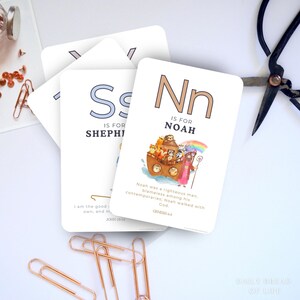 May include: White flashcards with the letters "S" and "N" printed on them. The "N" card features an illustration of Noah's Ark with animals and a rainbow. The text on the cards reads "IS FOR SHEPHERD" and "IS FOR NOAH".