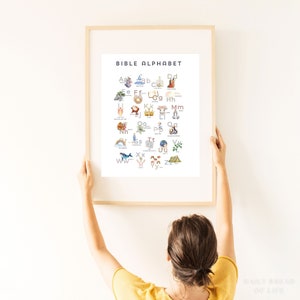 May include: A framed print titled "BIBLE ALPHABET" featuring watercolor illustrations for each letter. The artwork includes images of a whale, rainbow, and other religious symbols. The frame is light-colored wood, and the print is held by a person.