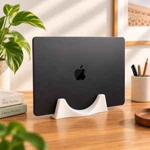 May include: A black laptop with a centered Apple logo is held upright by a white, curved, crescent-shaped stand. The stand is positioned on a wooden surface, providing a stable base for the device to remain in a vertical, upright orientation.