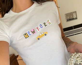 May include: A white t-shirt with a colourful "NEW YORK" graphic featuring a yellow taxi cab. The shirt is cropped and has a relaxed fit.