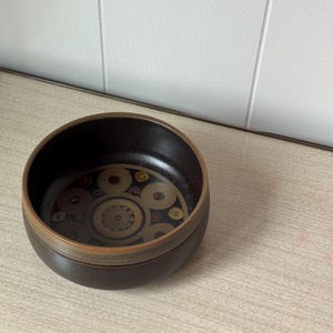 May include: A round, dark brown ceramic bowl with a decorative pattern in the center. The pattern features concentric circles and geometric shapes in shades of brown, tan, and gold. The rim of the bowl has a lighter brown band.