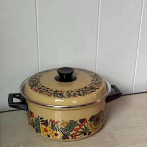 May include: A beige enamel pot with a lid, featuring a floral design in yellow, orange, green, and brown. The pot has black handles and a black knob on the lid. A vintage kitchenware item.