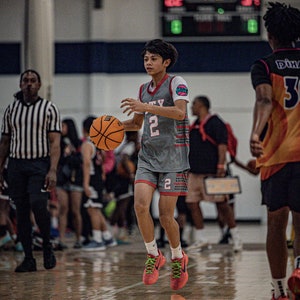 May include: A young basketball player wearing a gray jersey with the number 2 and the word "CITY" on it, dribbling a basketball. The player is wearing red and green sneakers and is on a basketball court.