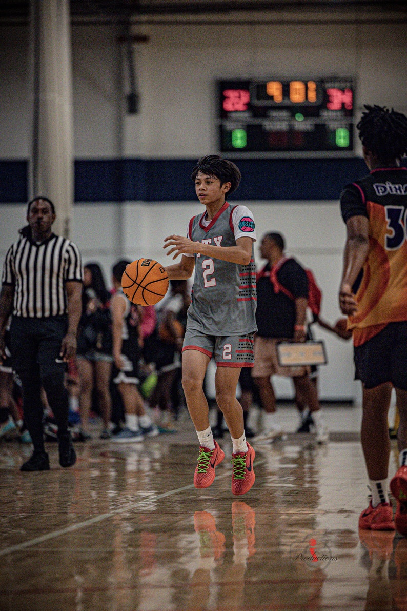 May include: A young basketball player wearing a gray jersey with the number 2 and the word "CITY" on it, dribbling a basketball. The player is wearing red and green sneakers and is on a basketball court.