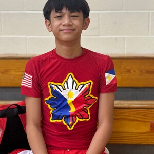 May include: A young person wearing a red shirt with a yellow star-shaped design featuring the Philippine flag. The shirt has a small American flag on the left sleeve and a Philippine flag on the right sleeve. The person is wearing a black wristband with the word "GRIT" on it.