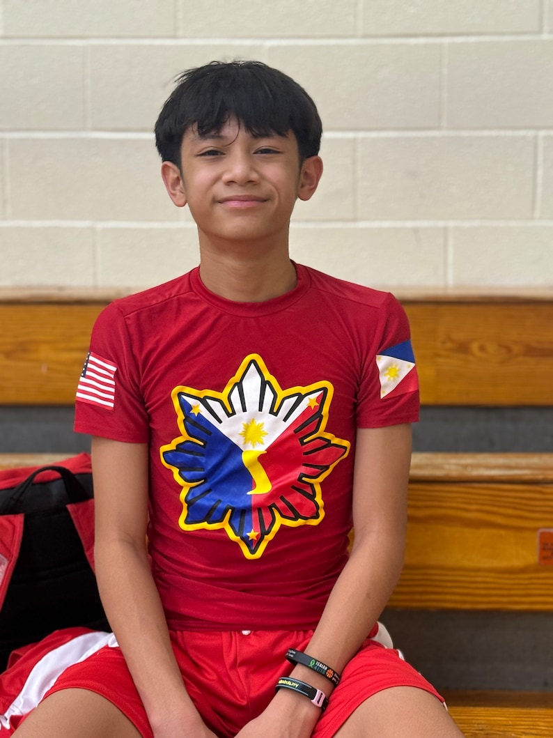 May include: A young person wearing a red shirt with a yellow star-shaped design featuring the Philippine flag. The shirt has a small American flag on the left sleeve and a Philippine flag on the right sleeve. The person is wearing a black wristband with the word "GRIT" on it.