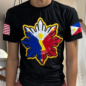May include: Black t-shirt with a yellow, red, blue, and white Philippine flag design on the front. The flag design features a sun with eight rays and a star in each corner. The sleeves have a small American flag on the left and a small Philippine flag on the right.