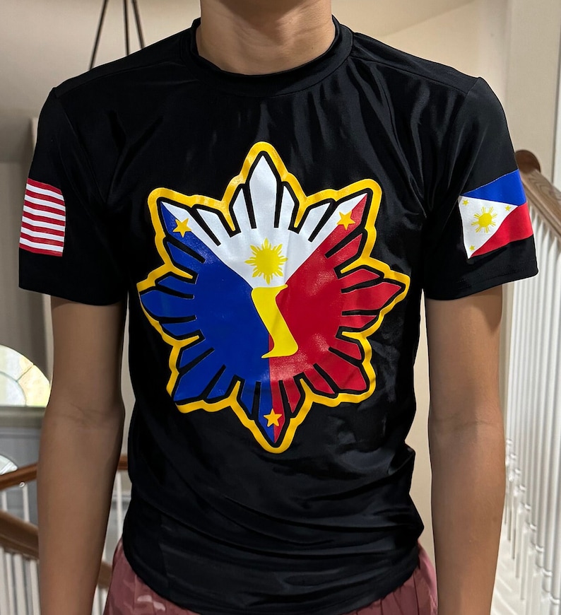 May include: Black t-shirt with a yellow, red, blue, and white Philippine flag design on the front. The flag design features a sun with eight rays and a star in each corner. The sleeves have a small American flag on the left and a small Philippine flag on the right.