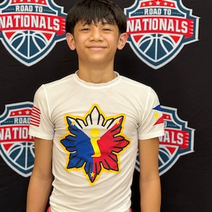 May include: A young boy wearing a white t-shirt with a large star-shaped graphic featuring the colors of the Philippine flag. The graphic has a yellow outline and a yellow number "1" in the center. The shirt has a small American flag on the left sleeve. The boy is standing in front of a black background with two basketball-themed signs that say "Road to Nationals".