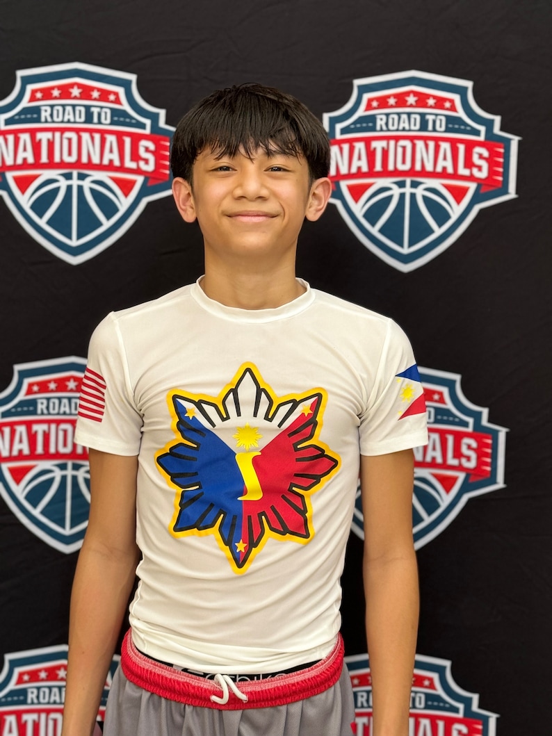 May include: A young boy wearing a white t-shirt with a large star-shaped graphic featuring the colors of the Philippine flag. The graphic has a yellow outline and a yellow number "1" in the center. The shirt has a small American flag on the left sleeve. The boy is standing in front of a black background with two basketball-themed signs that say "Road to Nationals".