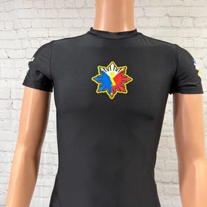 May include: Black short-sleeve shirt featuring a colorful emblem on the chest. The emblem is a sunburst design with red, white, blue, and yellow accents. The shirt is displayed on a mannequin.