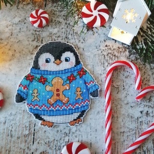 May include: A cross-stitched penguin wearing a blue sweater with a gingerbread man and floral design. The penguin is surrounded by red and white peppermint sweets, candy canes, and small white decorative houses with snowflake cutouts.