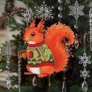 Könnte beinhalten: Ein Kreuzstich-Weihnachtsschmuck mit einem Eichhörnchen, das einen grünen Pullover und einen roten Kragen trägt und einen kleinen grünen Kiefernbaum hält. Der Schmuck ist orange, grün, rot und weiß. Der Hintergrund zeigt einen Weihnachtsbaum und dekorative Schneeflocken.