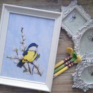 Puede incluir: Una bordadura de punto de cruz enmarcada en blanco con un pájaro azul y amarillo posado en una rama de flores blancas sobre un fondo azul claro.