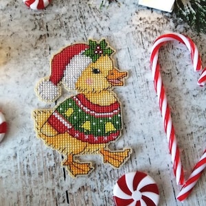 Può includere: Un'anatra gialla che indossa un maglione di Natale verde e rosso con campane e un cappello da Babbo Natale. L'anatra è in piedi su una superficie di legno bianco con bastoncini di zucchero e caramelle alla menta.