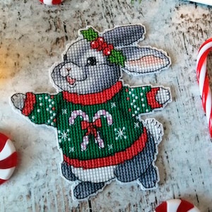 Puede incluir: Un conejito gris con un suéter navideño verde y rojo con bastones de caramelo y copos de nieve. El conejito tiene una rama de acebo en la cabeza.