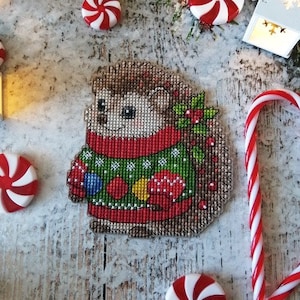 Puede incluir: Un patrón de punto de cruz de un erizo que lleva un suéter navideño verde y rojo con adornos. El erizo está rodeado de bastones de caramelo y caramelos de menta.