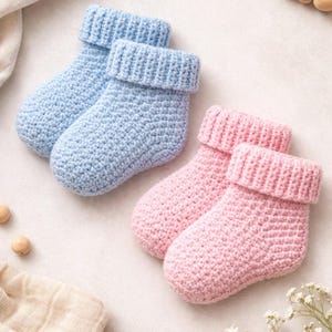 May include: Two pairs of baby socks, one pair in light blue and the other in pink, are displayed. The socks are handmade with a crochet stitch and have folded-over cuffs. The background includes wooden beads and a heart-shaped decoration.