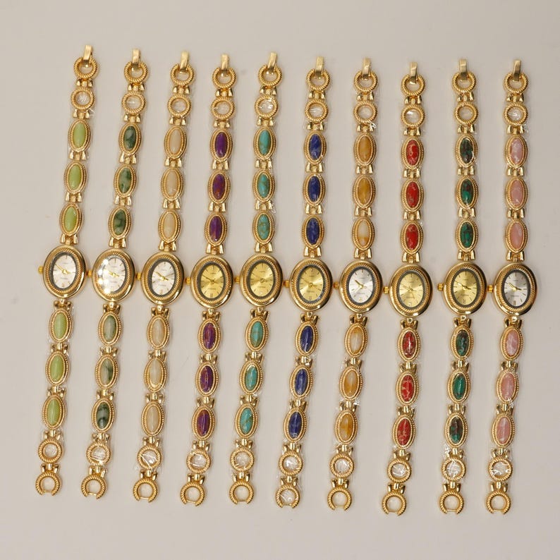 Puede incluir: Una colecci&oacute;n de relojes de pulsera dorados con esferas ovaladas y correas a juego. Cada correa presenta una serie de piedras preciosas ovaladas de colores, incluyendo verde, morado, azul y rojo. Las esferas de los relojes son doradas o plateadas.