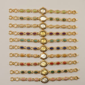 Puede incluir: Una colecci&oacute;n de relojes de pulsera dorados con esferas ovaladas y correas adornadas con piedras preciosas de colores. Cada reloj presenta un color de piedra diferente, incluyendo verde, morado, azul y rosa. Los relojes se exhiben sobre un fondo claro.