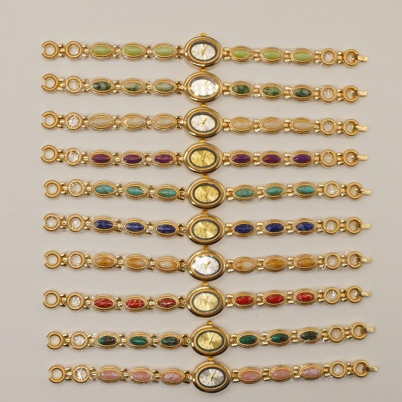 Puede incluir: Una colecci&oacute;n de relojes de pulsera dorados con esferas ovaladas y correas adornadas con piedras preciosas de colores. Cada reloj presenta un color de piedra diferente, incluyendo verde, morado, azul y rosa. Los relojes se exhiben sobre un fondo claro.