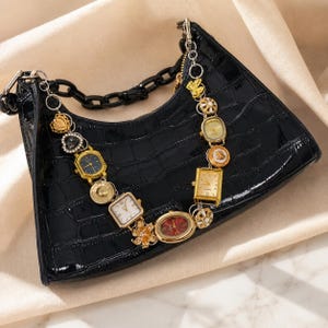 Puede incluir: Bolso de mano negro con efecto cocodrilo y correa de reloj. La correa está compuesta por varias esferas de reloj doradas y joya. El bolso tiene un asa de cadena negra y un cierre plateado. Aproximadamente 25 cm de ancho.