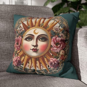 May include: A square throw pillow in a deep teal color featuring a central illustration of a sun face with a crescent moon on its forehead. The sun is surrounded by golden rays and pink floral arrangements, all enclosed within an ornate frame.