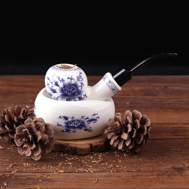 May include: A white ceramic pipe with blue floral designs sits in a matching ceramic holder. The pipe has a black stem and is filled with tobacco. The pipe and holder are on a wooden surface with pine cones.